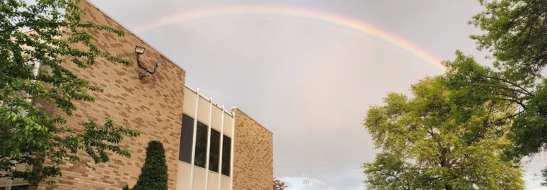 A rainbow arches over a brick building, framed by green trees.