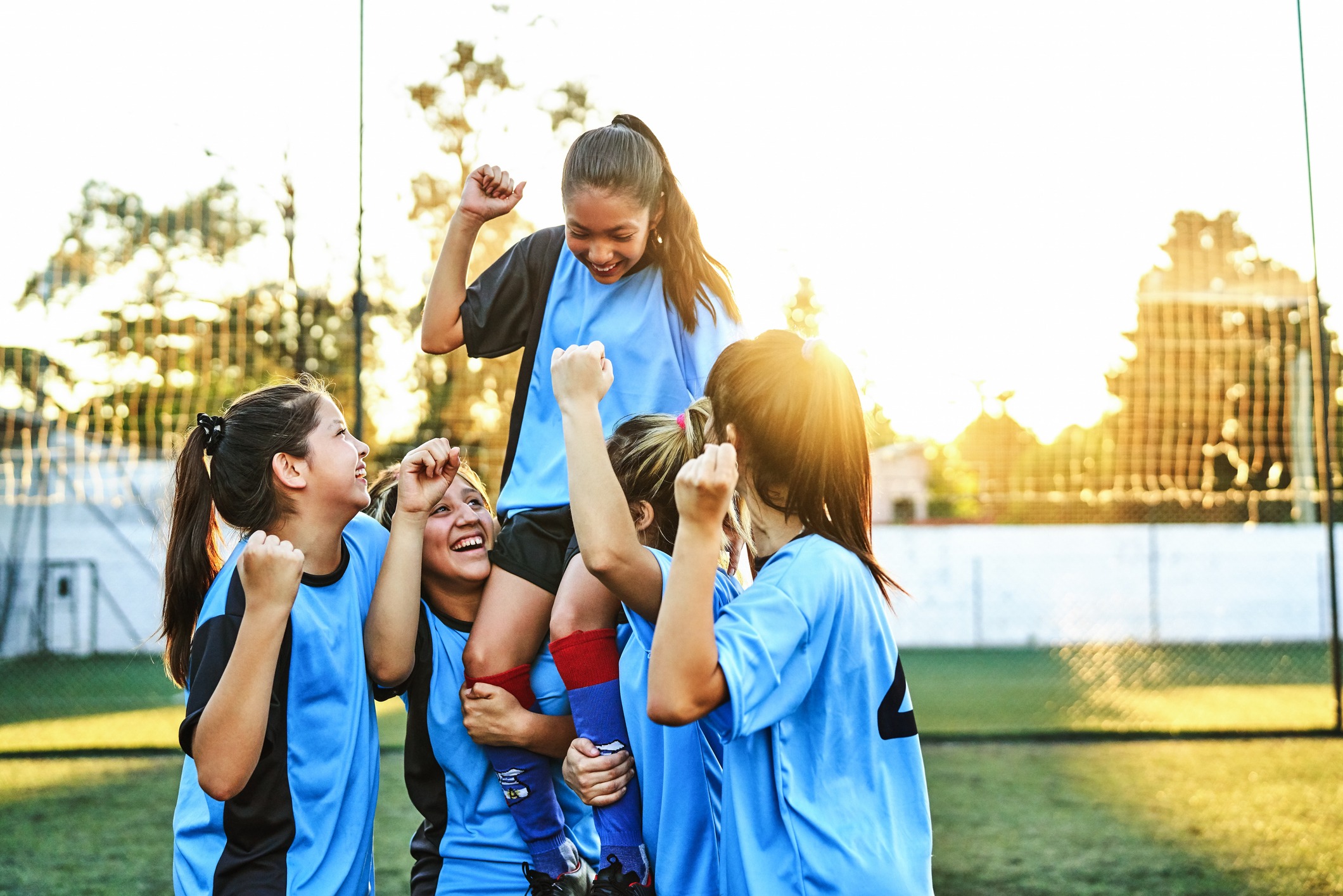 A group of young women in blue jerseys celebrate on a soccer field, lifting a teammate.