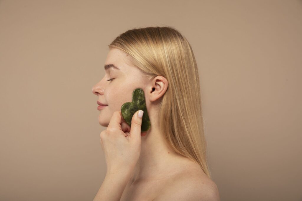 A person using a green jade gua sha tool on their cheek, eyes closed, against a beige background.