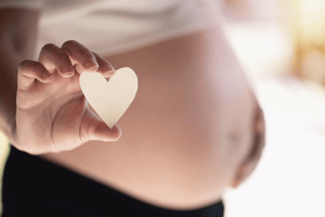 A hand holds a small paper heart in front of a pregnant belly.