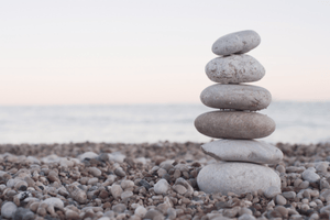 Stack of smooth stones balanced on a pebble-covered beach with a calm sea and pastel horizon in the background.